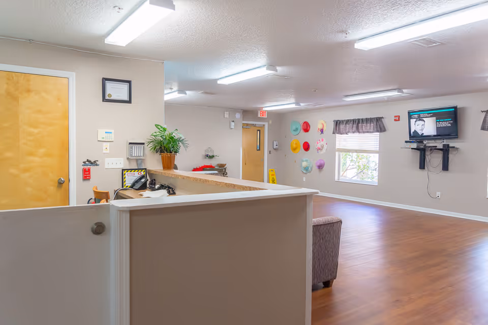 Interior view of a senior living facility reception area with a wooden door, a desk with a plant and telephone, a wall decorated with colorful hats, two windows with curtains, a wall-mounted TV, and wooden flooring.