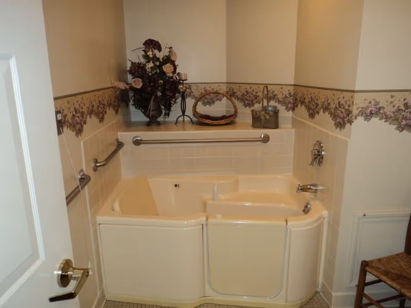 Walk-in bathtub with a safety door and grab bars, floral wallpaper border, and decorative items on a tiled ledge in a bathroom.