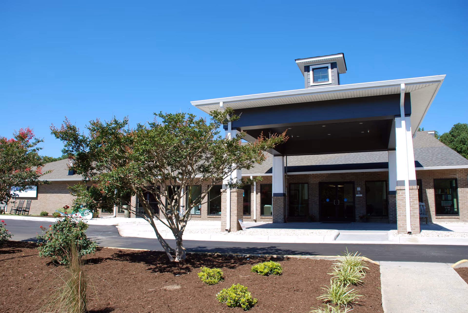 Front entrance of a single-story brick building with a covered porte-cochère and landscaped beds.