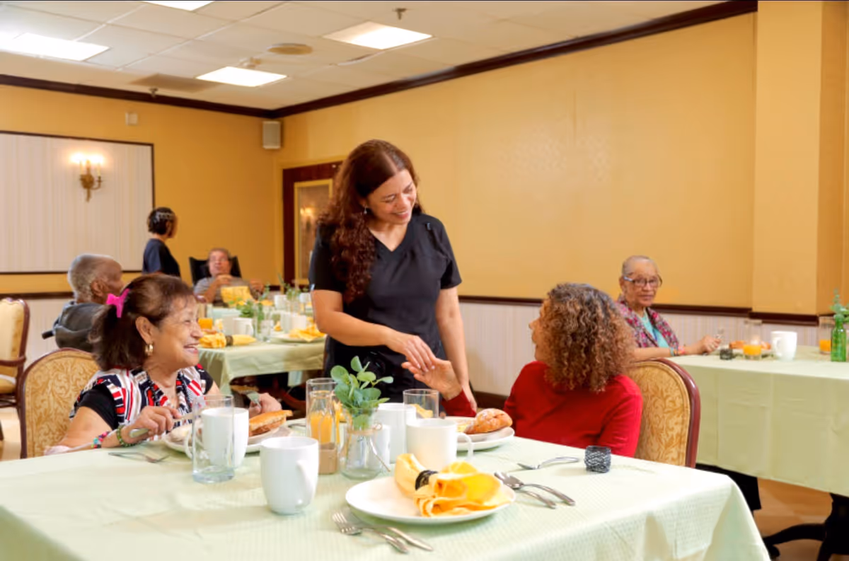 A dining room in a senior living facility where elderly residents are seated at tables covered with light green tablecloths, enjoying a meal. A staff member in black scrubs is smiling and interacting warmly with two women seated at the front table. The room has yellow walls and soft lighting.