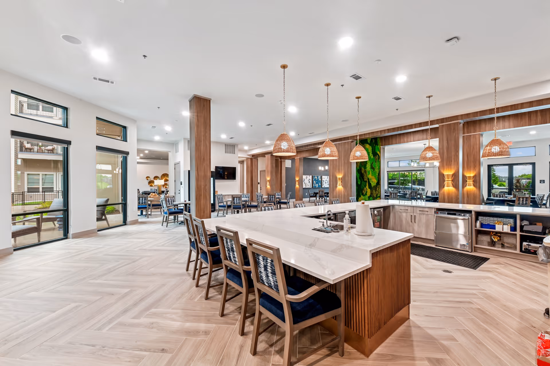 A spacious, modern dining area with a large white marble island countertop surrounded by wooden chairs with blue cushions. The room features wooden columns, hanging wicker pendant lights, and large windows letting in natural light. There are additional dining tables and chairs in the background, and a green moss wall decoration is visible.