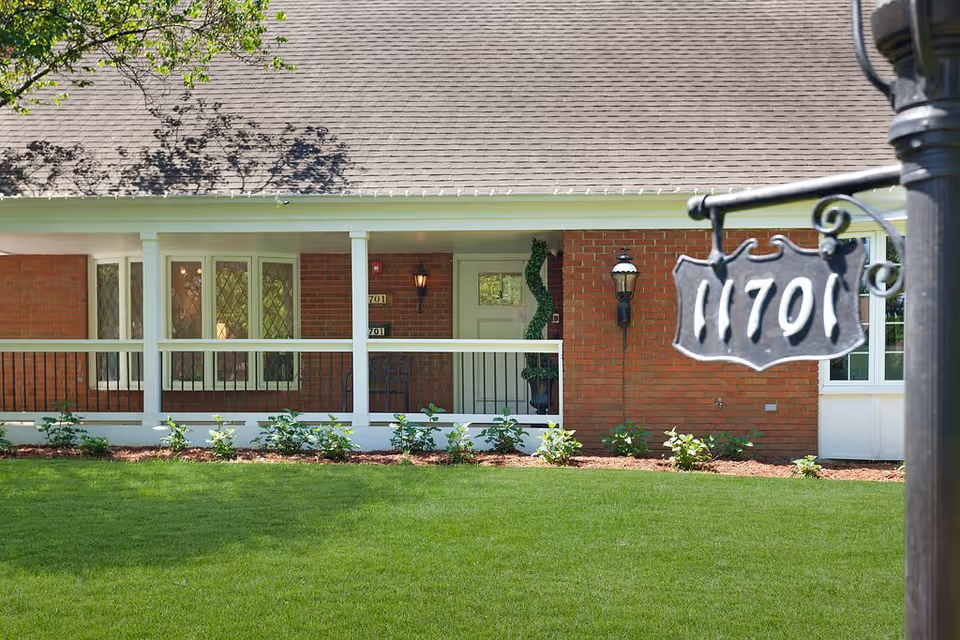Front exterior view of a single-story brick building with a covered porch, white railing, and a green lawn in front. The building has a door with the number 701 and windows with decorative glass. A black metal signpost with the number 1701 is visible in the foreground.
