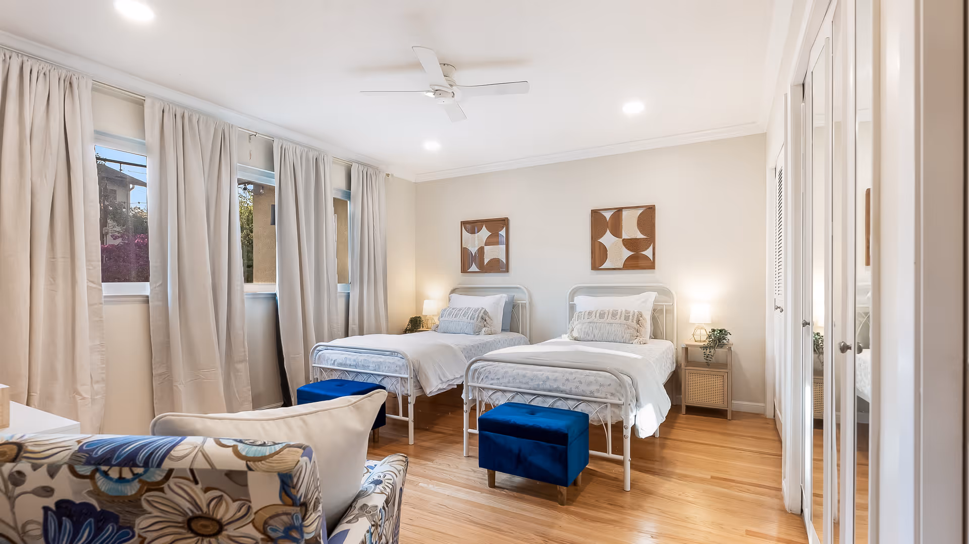 Bright bedroom with two twin beds, blue ottomans, an armchair, and large curtained windows.