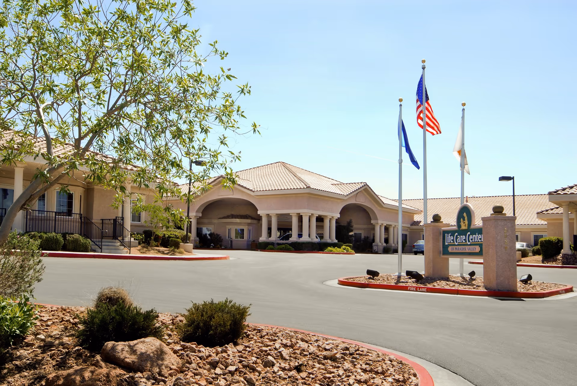 Front exterior view of the Life Care Center of South Las Vegas building with a circular driveway, three flagpoles displaying flags, desert landscaping with rocks and bushes, and a clear blue sky.