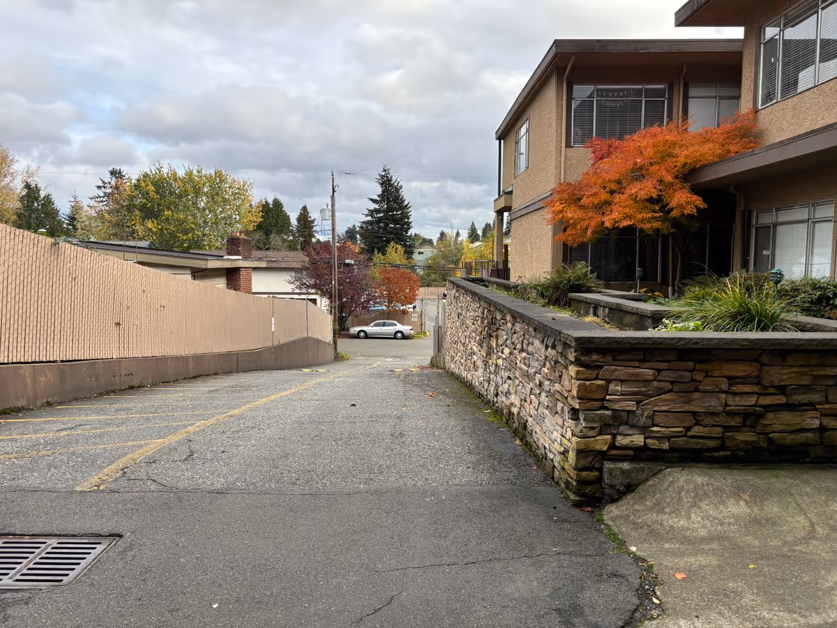 Driveway and parking area beside a two-story building with a stone retaining wall and an orange autumn tree under a cloudy sky.