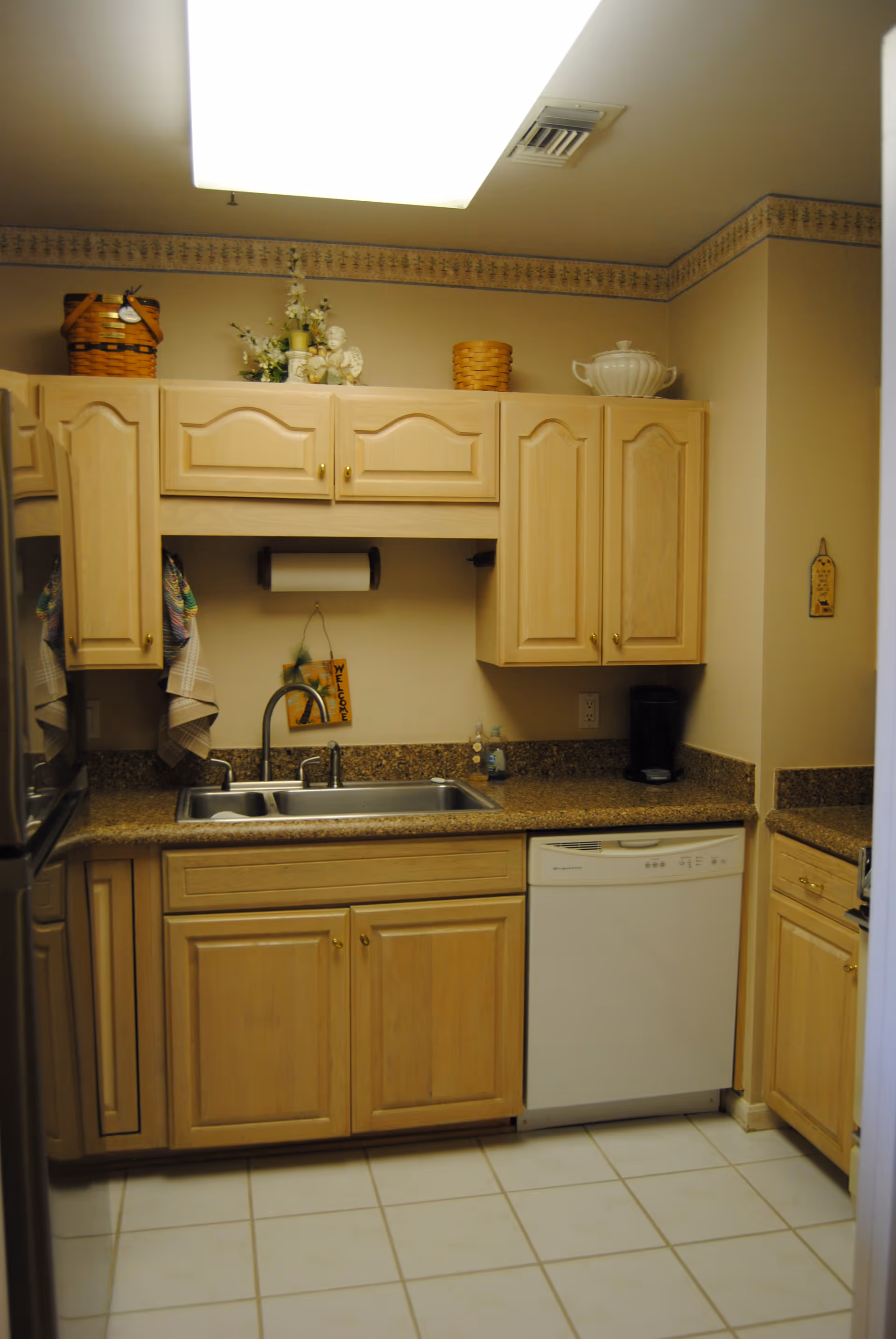 A small kitchen area with light wood cabinets, a double stainless steel sink, a white dishwasher, and a granite countertop. There are decorative baskets and a white ceramic dish on top of the cabinets. The walls are beige with a floral border near the ceiling, and the floor is tiled in white.