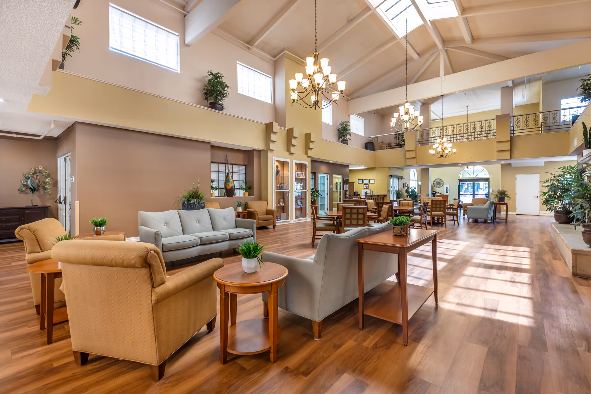 Spacious senior living facility common area with high vaulted ceilings, large windows, and multiple seating arrangements including sofas, armchairs, and wooden tables. The room features warm wood flooring, chandeliers, and decorative plants placed throughout the space.