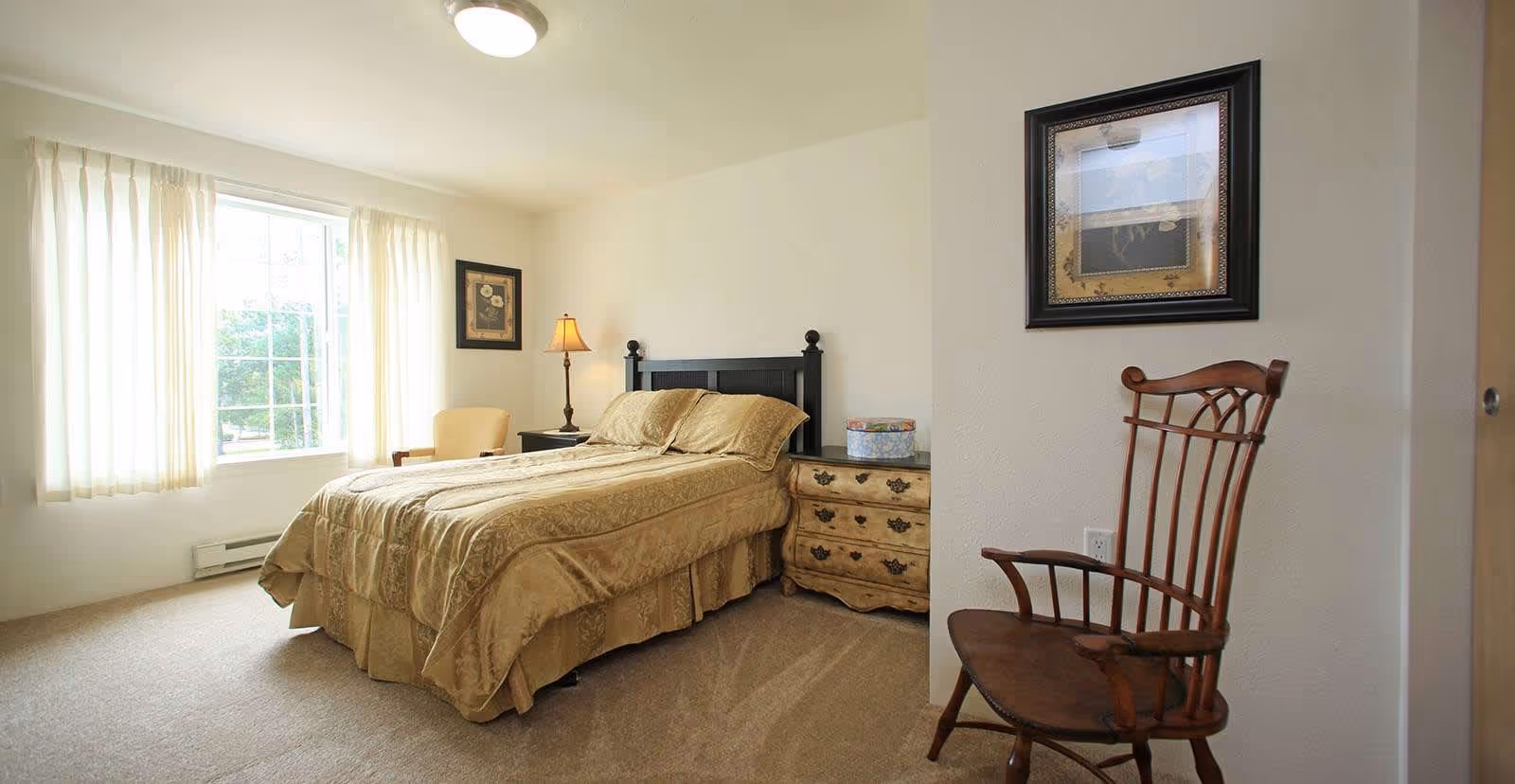 A bedroom with a large window covered by sheer curtains letting in natural light. The room features a bed with a beige comforter, a wooden headboard, a nightstand with a lamp, a framed picture on the wall, a wooden chair, and a carpeted floor.