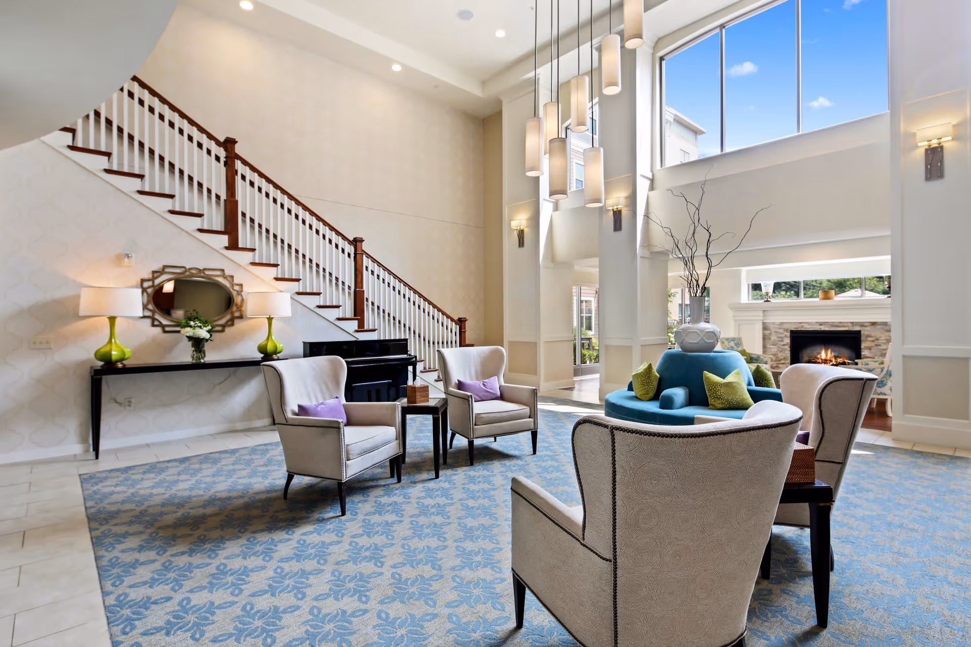 A spacious and well-lit living room area with high ceilings and large windows. The room features a blue patterned carpet, several beige armchairs with purple cushions arranged around a circular blue sofa with green pillows, a fireplace with a stone surround, a black piano, a console table with two green lamps and a decorative mirror, and a staircase with wooden handrails.