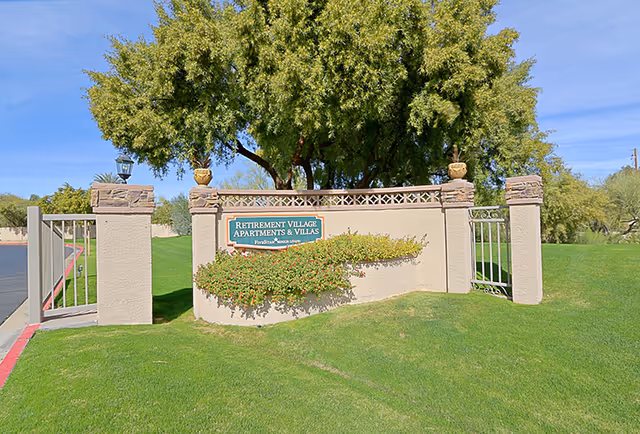 Entrance sign for a retirement village apartments and villas with a beige stone wall, green shrubbery, and a large tree in the background under a clear blue sky.