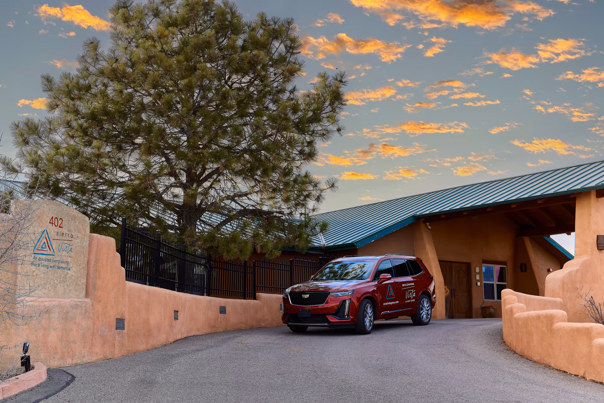 Exterior view of Sierra Vista at Vista Living Care facility during sunset, showing a red SUV parked on a driveway next to a building with a green metal roof and adobe-style walls. A large tree is visible behind a fence, and a sign on the wall displays the facility's name and address.