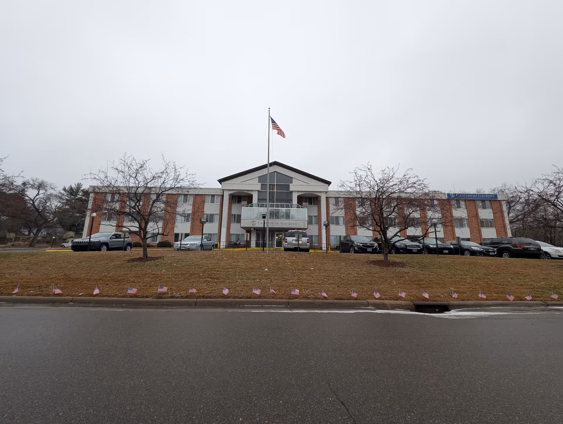 Front exterior view of Centennial Gardens for Nursing and Rehabilitation, a three-story building with a central entrance, an American flag on a flagpole in front, several parked cars, leafless trees, and small American flags lining the grass near the curb on a cloudy day.