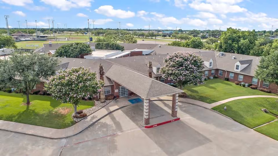 Aerial view of Morada Abilene senior living facility showing a single-story brick building with a covered entrance driveway, surrounded by green lawns, trees with pink blossoms, and a clear blue sky with scattered clouds.
