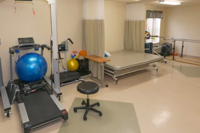 A rehabilitation room with a treadmill, exercise balls, a stool, a therapy bed with privacy curtains, parallel bars for walking exercises, and various therapy equipment in a well-lit room with beige walls and tiled floor.