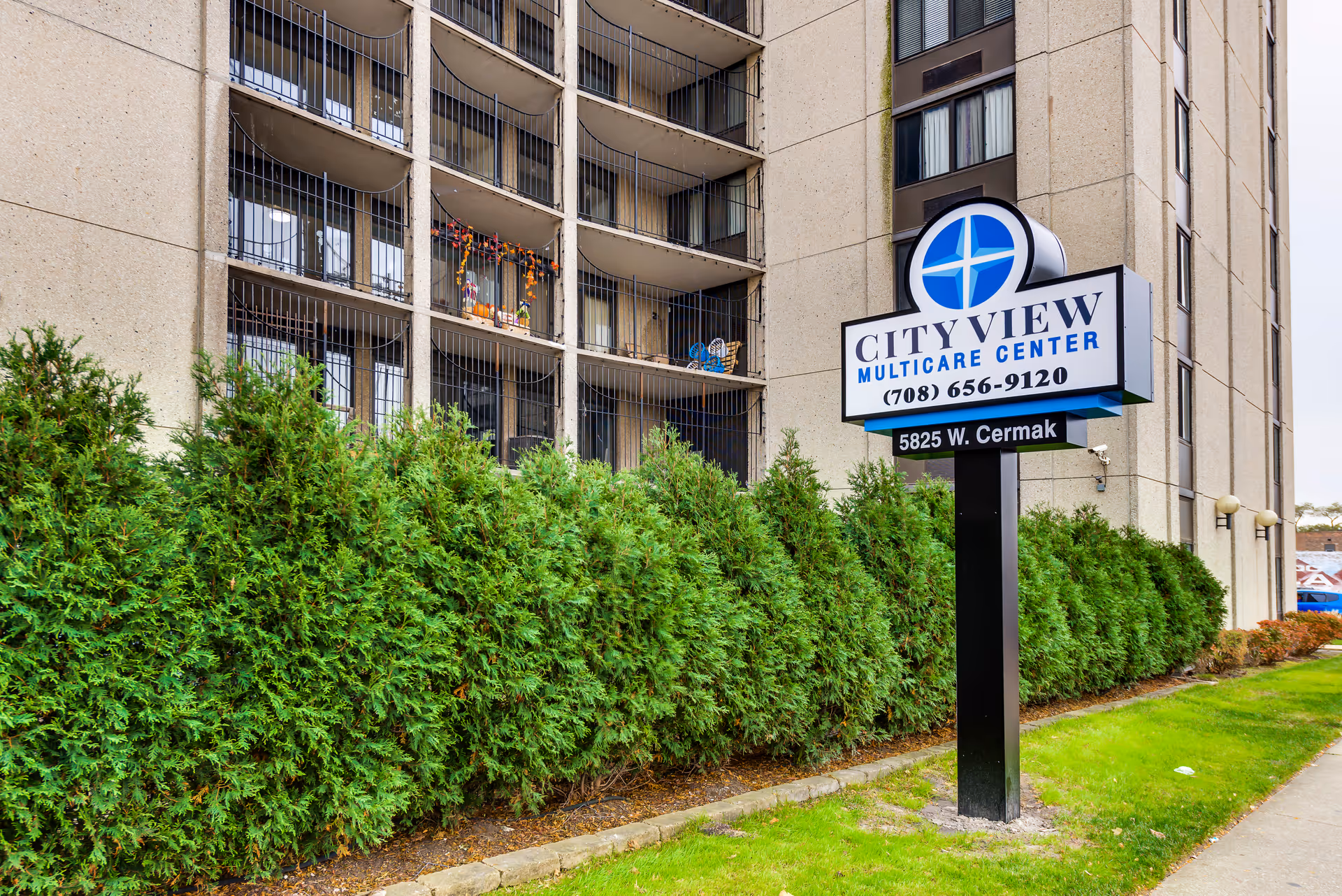 Exterior view of City View Multicare Center building with a sign displaying the facility name, phone number, and address. The building has multiple balconies with railings and some decorations visible. There are green bushes and a grassy area in front of the building.