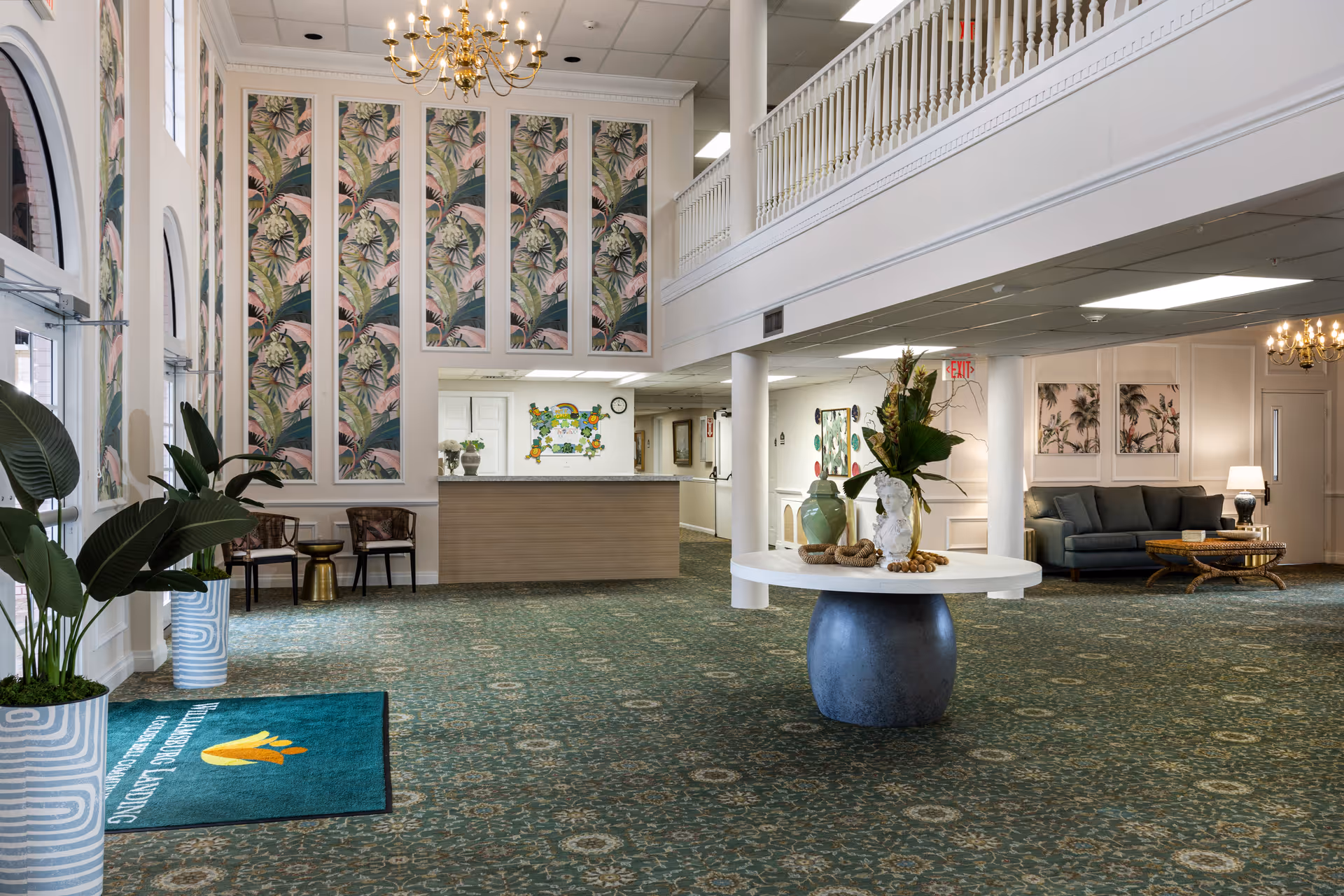 A spacious, well-lit assisted living lobby with large patterned wall panels, a central round table with decorative vases, seating areas, and a reception desk.
