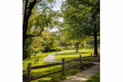 A peaceful outdoor scene at The Village at St. Barnabas featuring a winding paved pathway through a grassy area with several trees providing shade. A wooden fence runs alongside the path, and a bench is visible near the path under the trees.