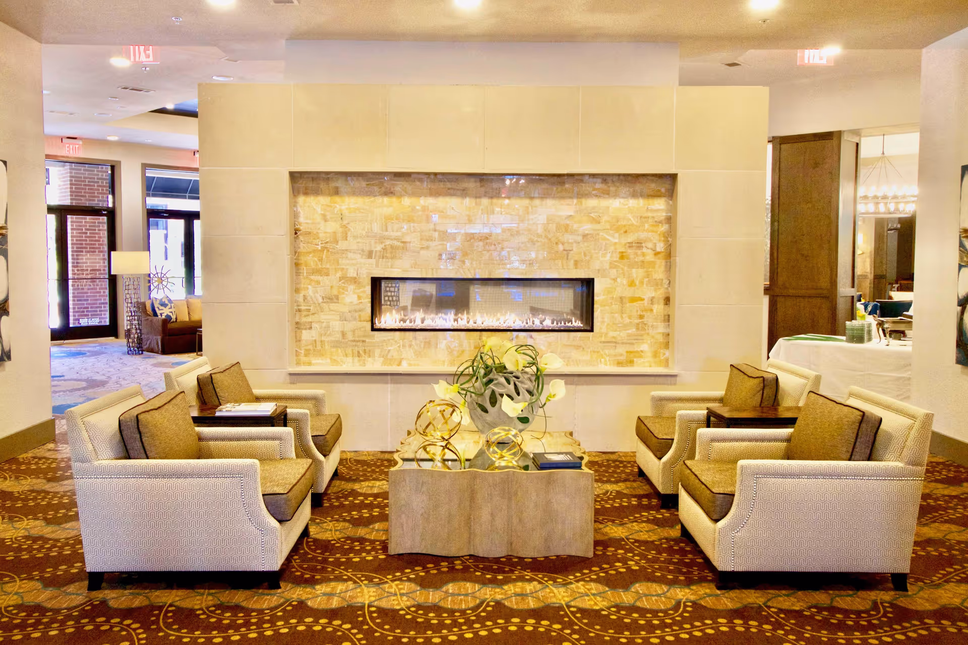 A cozy seating area in a senior living facility with four cushioned armchairs arranged around a small wooden coffee table. The table has decorative items including a plant and golden spherical sculptures. Behind the seating area is a modern rectangular gas fireplace set into a tiled wall. The room has warm lighting and a patterned carpet, with visible doorways leading to other parts of the facility.