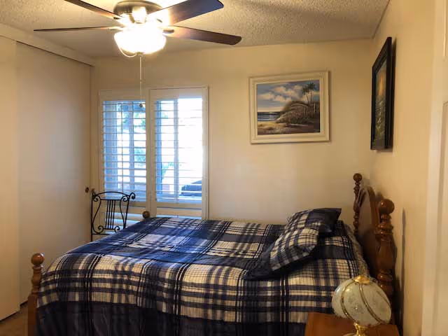 Small bedroom with a wooden bed covered in blue plaid bedding, a ceiling fan, a shuttered window, and framed artwork on the walls.
