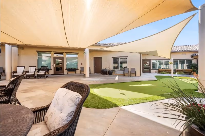 Outdoor courtyard area with a putting green, shaded by large beige triangular sun sails. There are wicker chairs with cushions and a table in the foreground, and several more chairs lined up against the building walls. The building has large windows and doors opening to the courtyard under a clear blue sky.