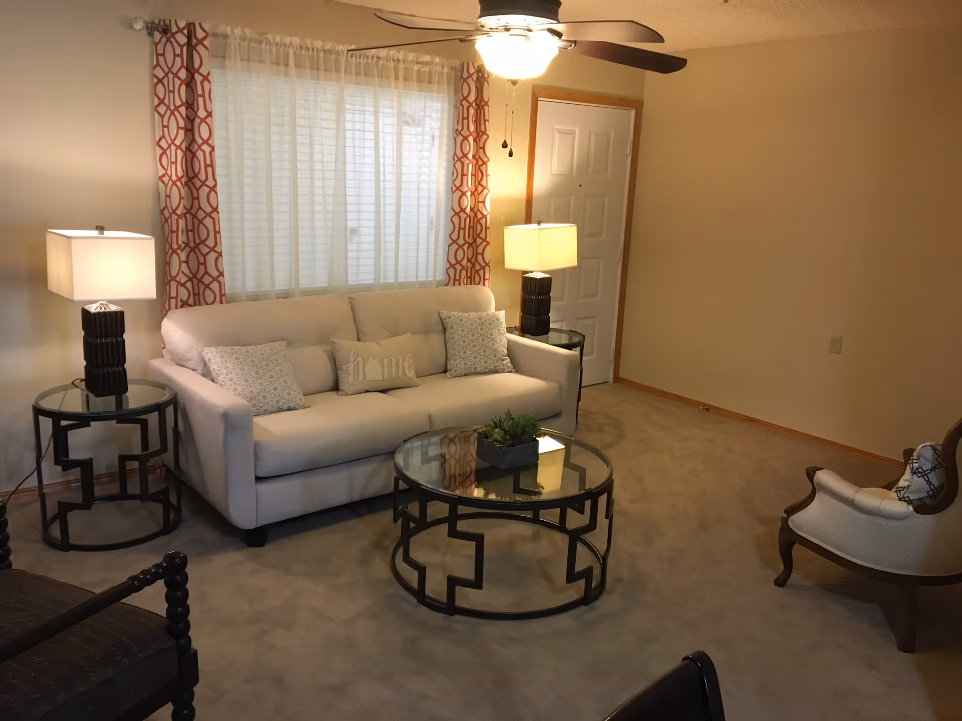 A cozy living room with a beige sofa adorned with three decorative pillows, two side tables each with a lamp, a round glass coffee table with a small plant centerpiece, a window with white blinds and orange patterned curtains, a ceiling fan with a light, and an armchair with a patterned pillow.