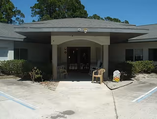 Entrance to a single-story building with a covered porch area supported by columns. There are two plastic chairs placed near the entrance and some bushes along the front of the building. The sky is clear and blue.