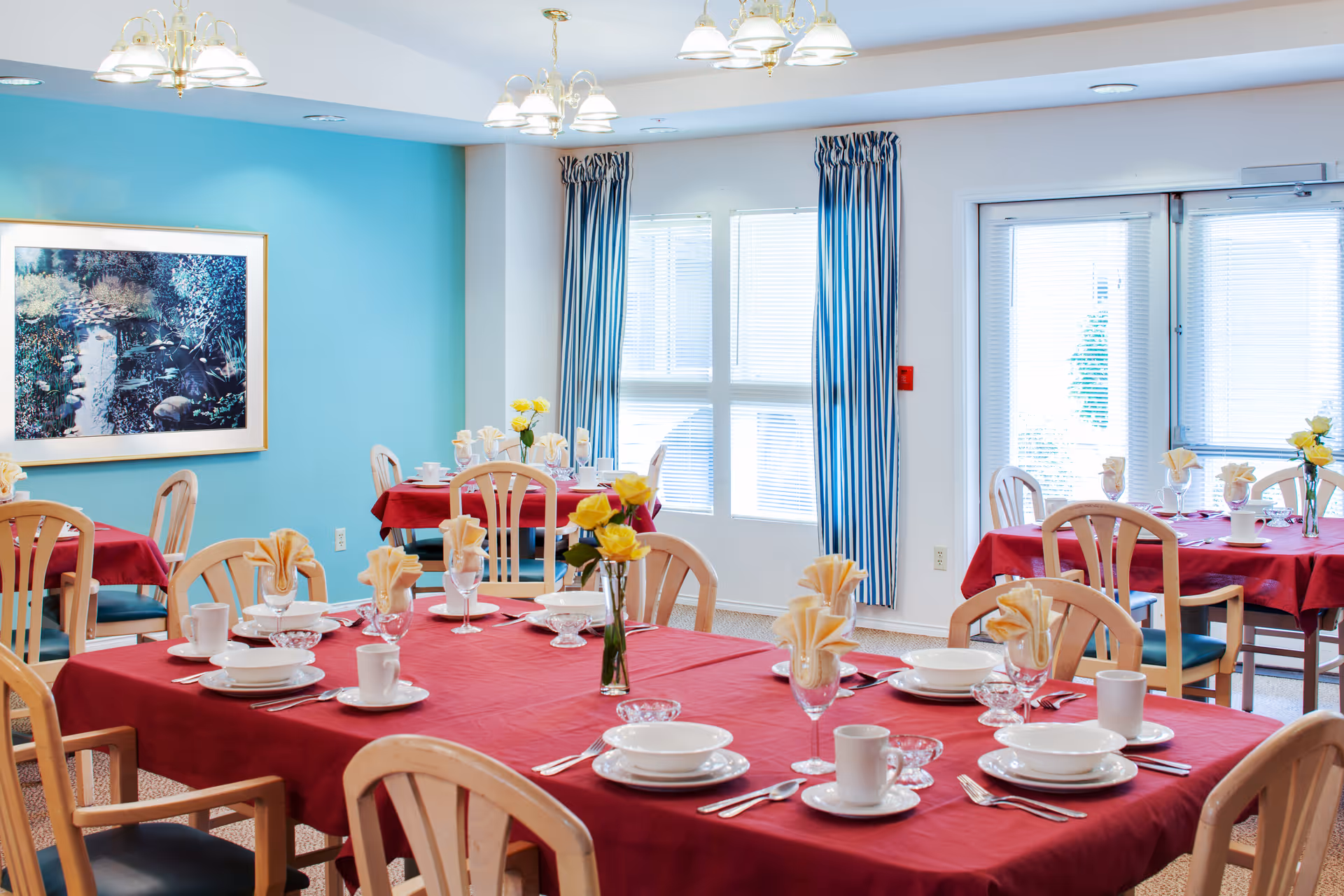 Bright dining room with multiple tables covered in red tablecloths, set with plates, cups, cutlery and yellow flower centerpieces.