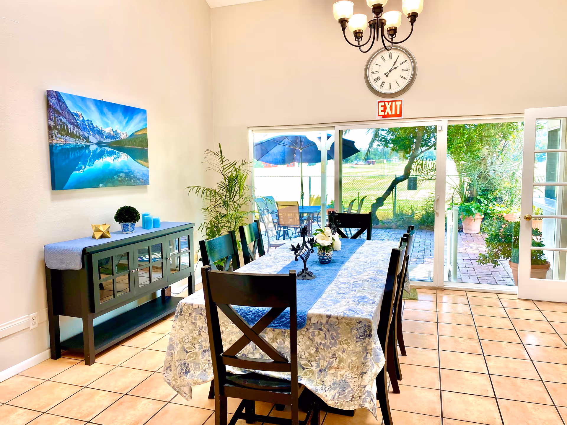 A bright dining room with a long table covered with a floral tablecloth and a blue runner, surrounded by black wooden chairs. A sideboard with decorative items and a landscape painting is against the wall. Large sliding glass doors open to an outdoor patio with tables, chairs, and umbrellas. A clock and an exit sign are mounted above the doors.