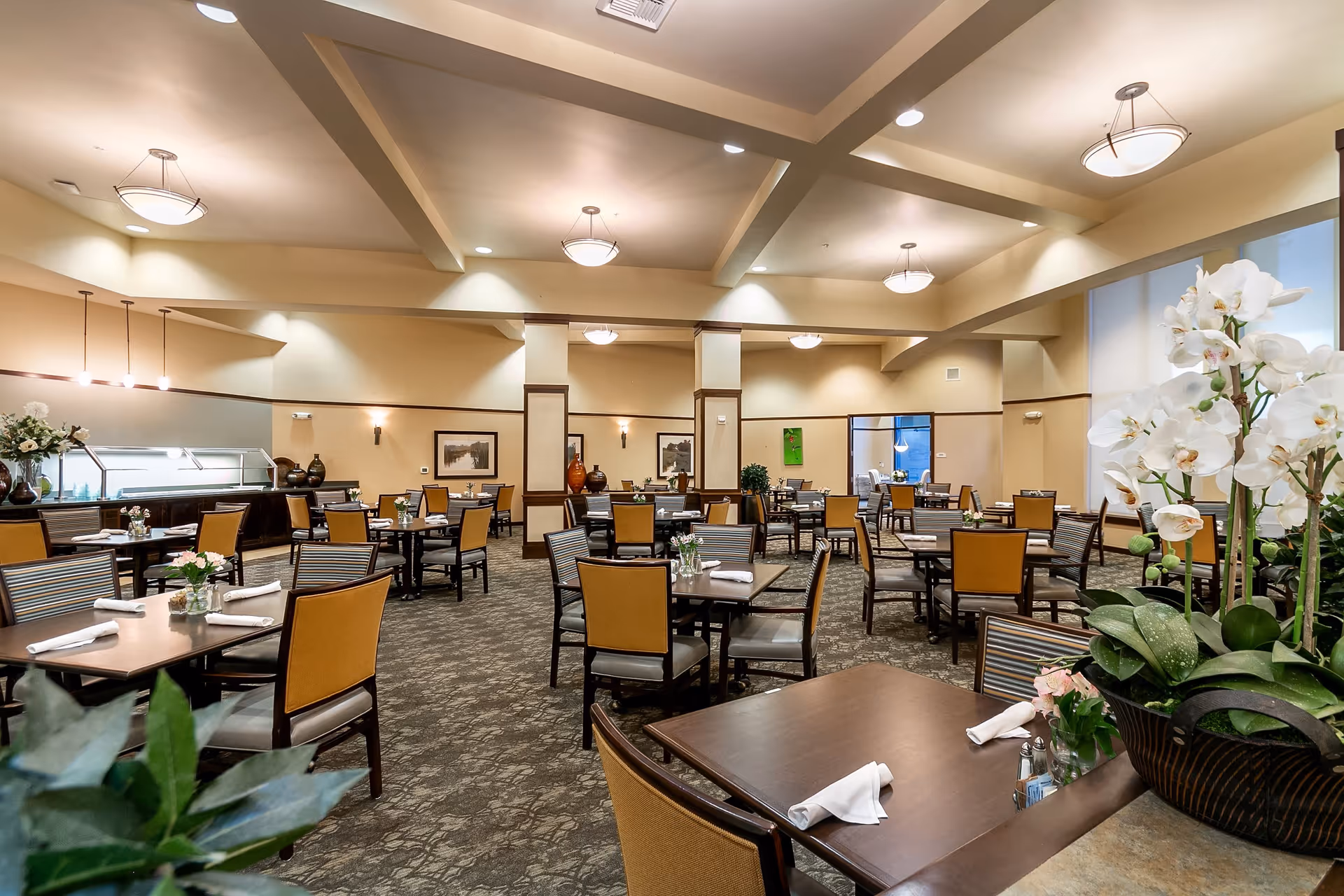 A spacious dining room with multiple tables and chairs arranged neatly. Each table is set with rolled white napkins and small flower arrangements. The room has a high ceiling with recessed lighting and pendant lights. There are decorative vases and framed pictures on the walls, and a large floral arrangement is visible in the foreground.
