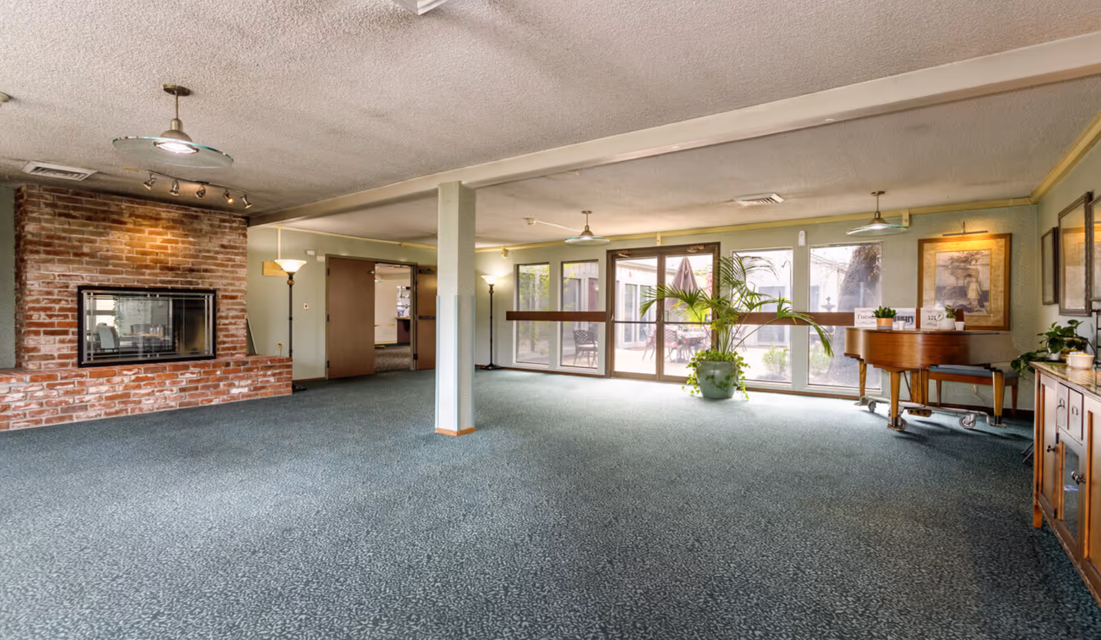 Spacious carpeted common room with a brick fireplace, large windows overlooking a patio, potted plants, and a piano.