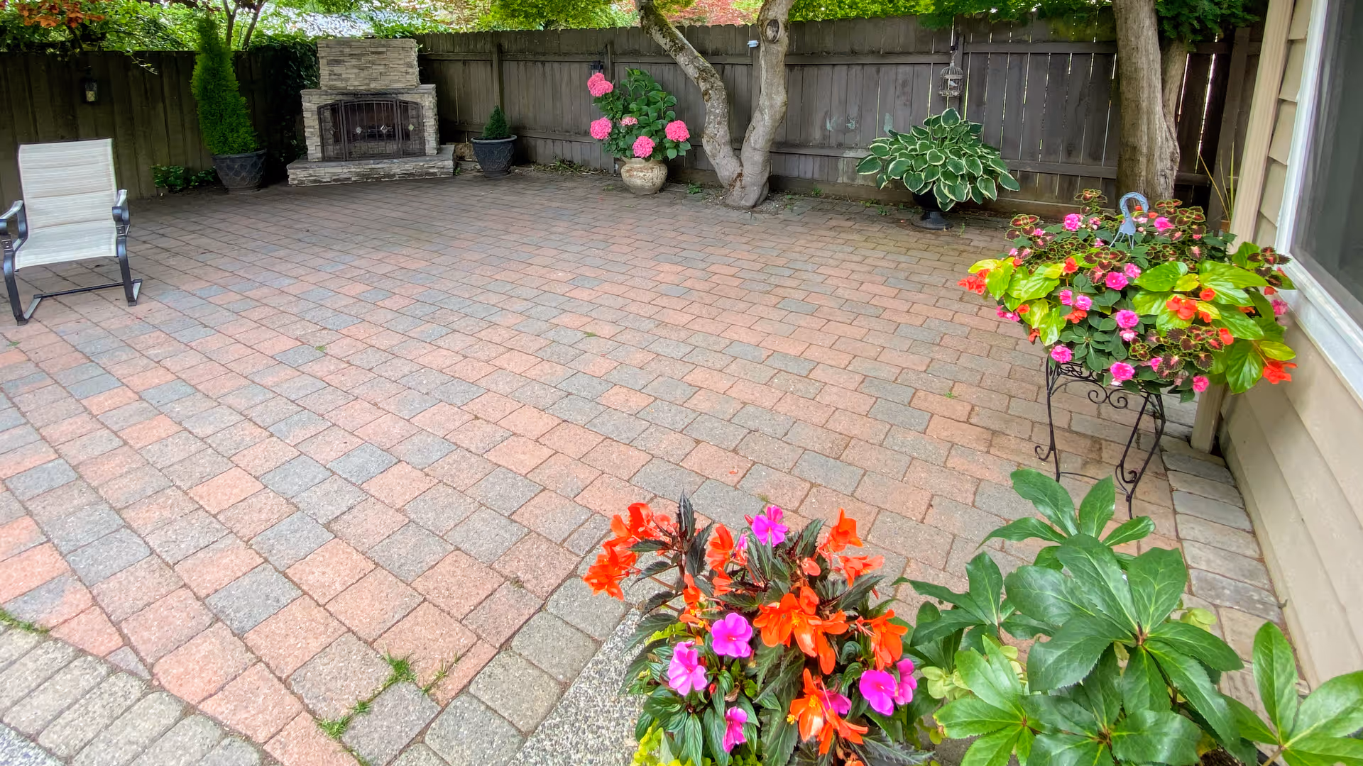 A spacious outdoor patio area with a tiled floor, surrounded by a wooden fence. There is a stone fireplace against the fence, several potted plants with colorful flowers, and a single chair on the left side. Trees and greenery are visible around the patio.
