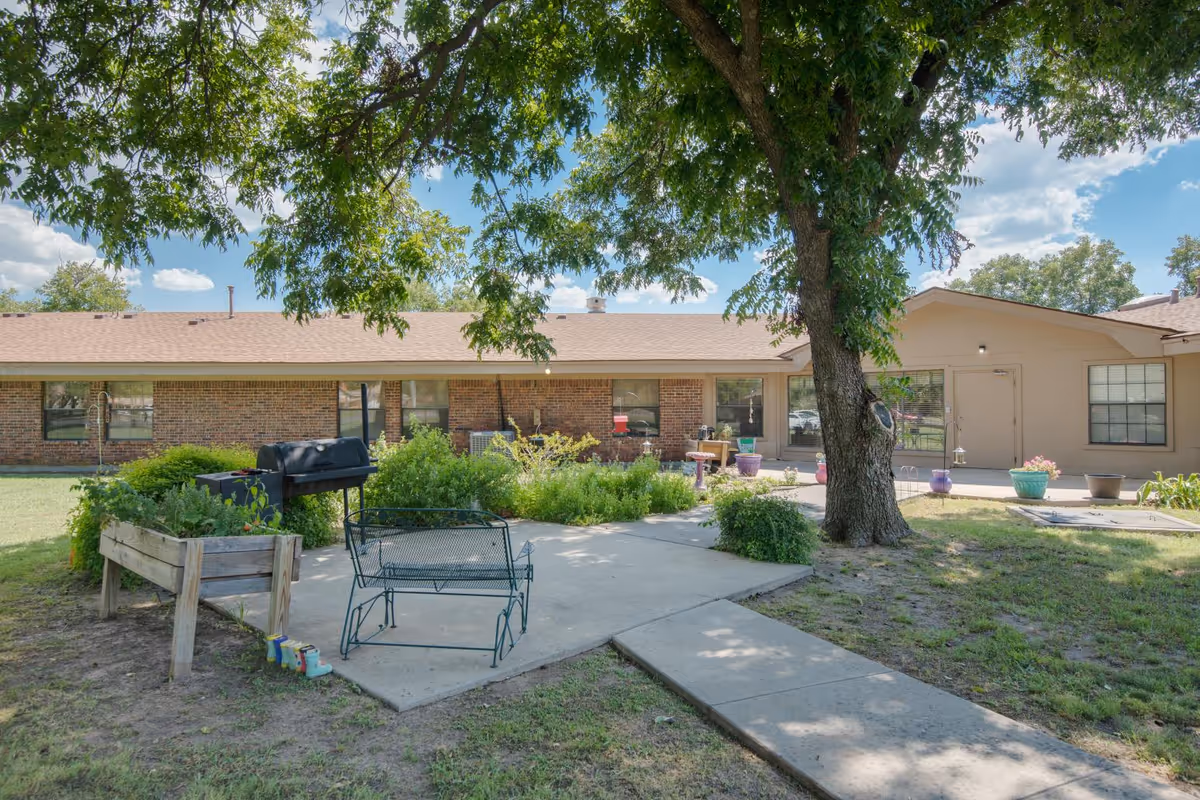 Outdoor patio area at Pecan Tree Rehab and Healthcare Center featuring a large tree providing shade, a metal bench, a barbecue grill, raised garden beds with plants, and a concrete walkway leading to the building entrance. The building has brick and beige siding with several windows and potted plants near the door.