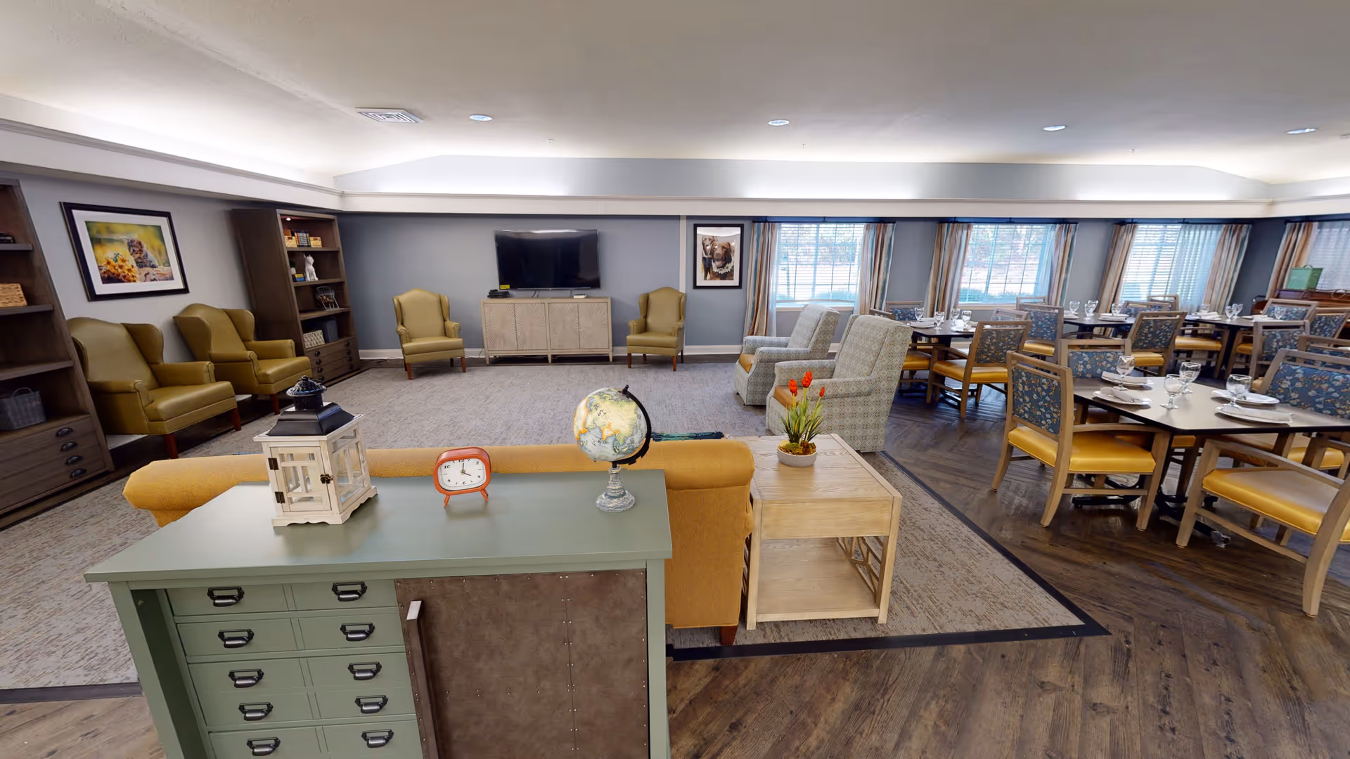 Spacious communal lounge and dining area with sofas and armchairs facing a television on the left and multiple set dining tables on the right.