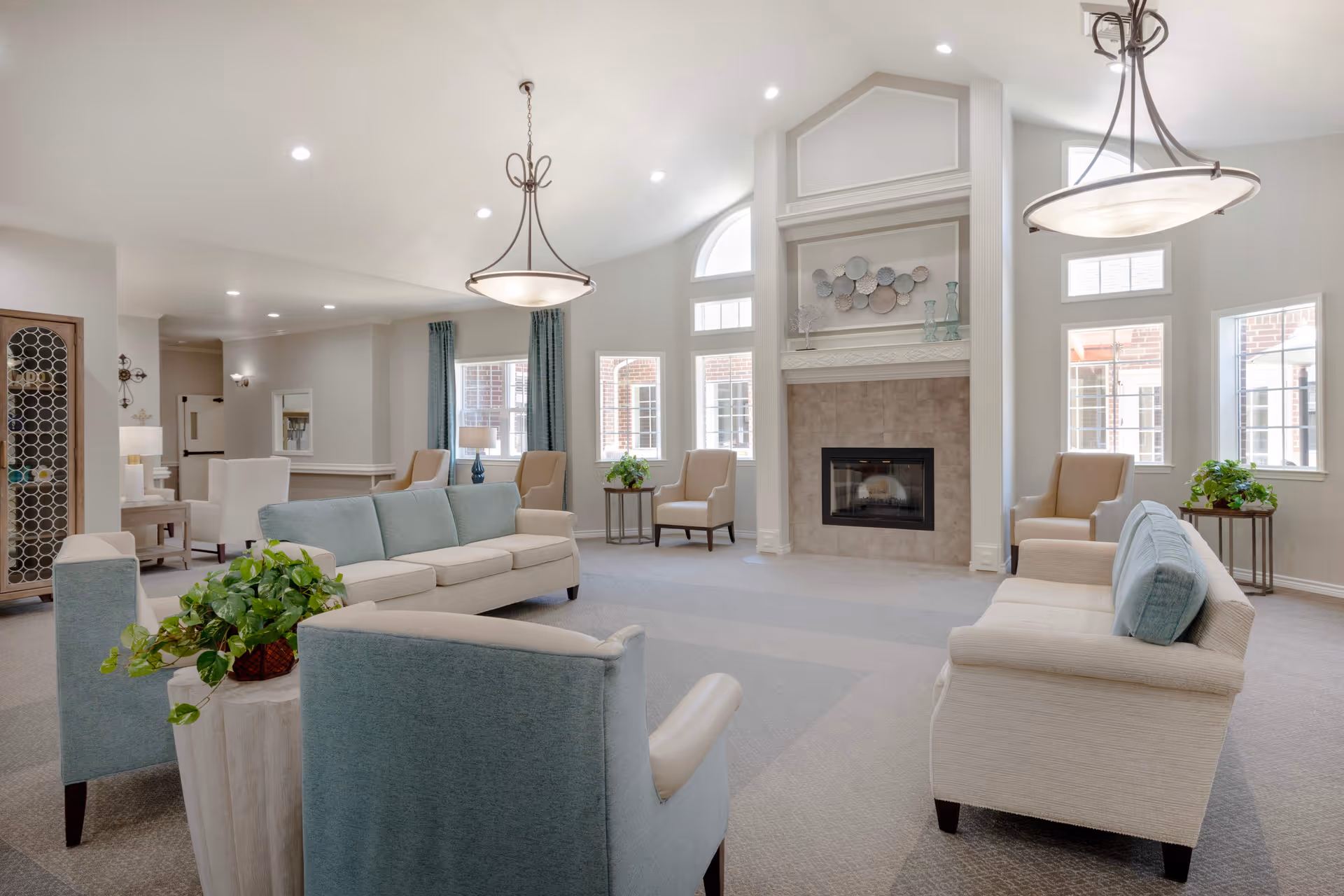 A bright and spacious living room area in Oak Ridge Alzheimer's Special Care Center featuring multiple beige and light blue upholstered sofas and armchairs arranged around a central fireplace with decorative art above it. The room has large windows allowing natural light, two hanging light fixtures, and several small tables with green plants.