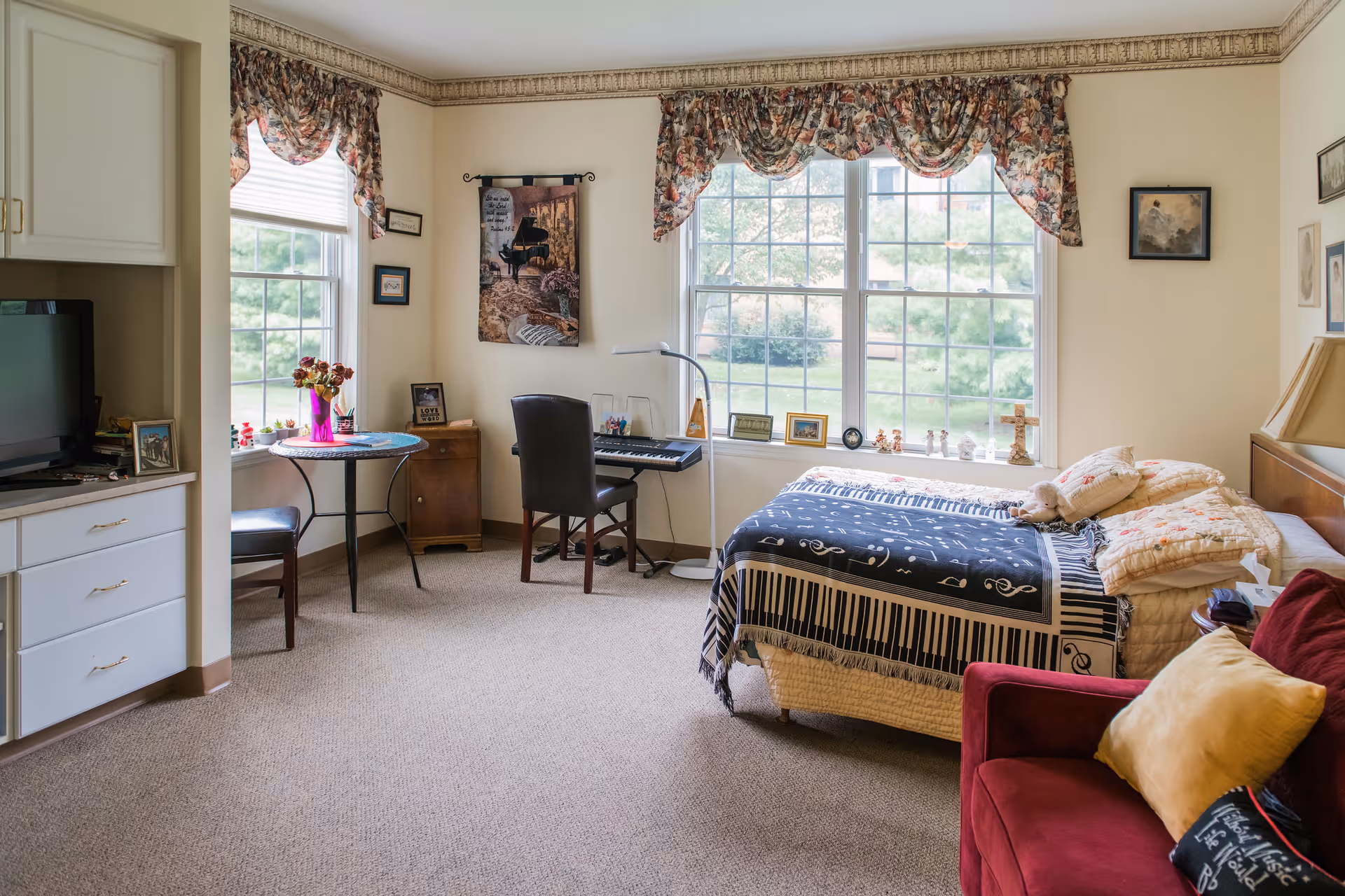 A cozy bedroom with a bed covered in a musical notes and piano keys themed blanket, a red armchair with yellow pillows, a small round table with a vase of flowers, a chair, and a keyboard piano by the window. The room has large windows with floral valances, beige walls, and carpeted floor. Various framed pictures and decorations are on the walls and windowsill.
