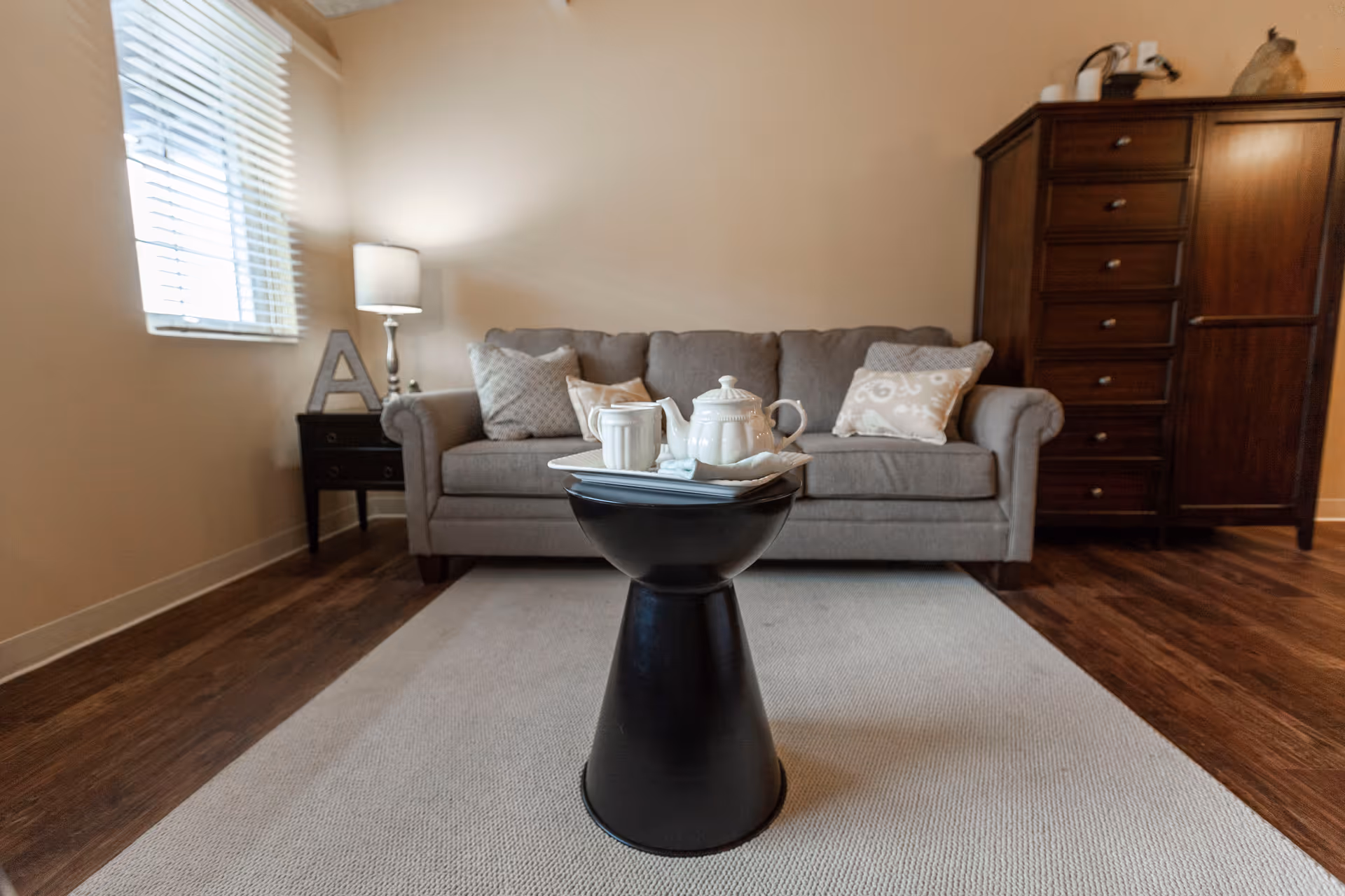 A cozy living room with a gray sofa adorned with patterned cushions, a black modern side table in front holding a white teapot and cup on a tray, a wooden dresser to the right, and a small side table with a lamp and decorative letter 'A' near a window with blinds.