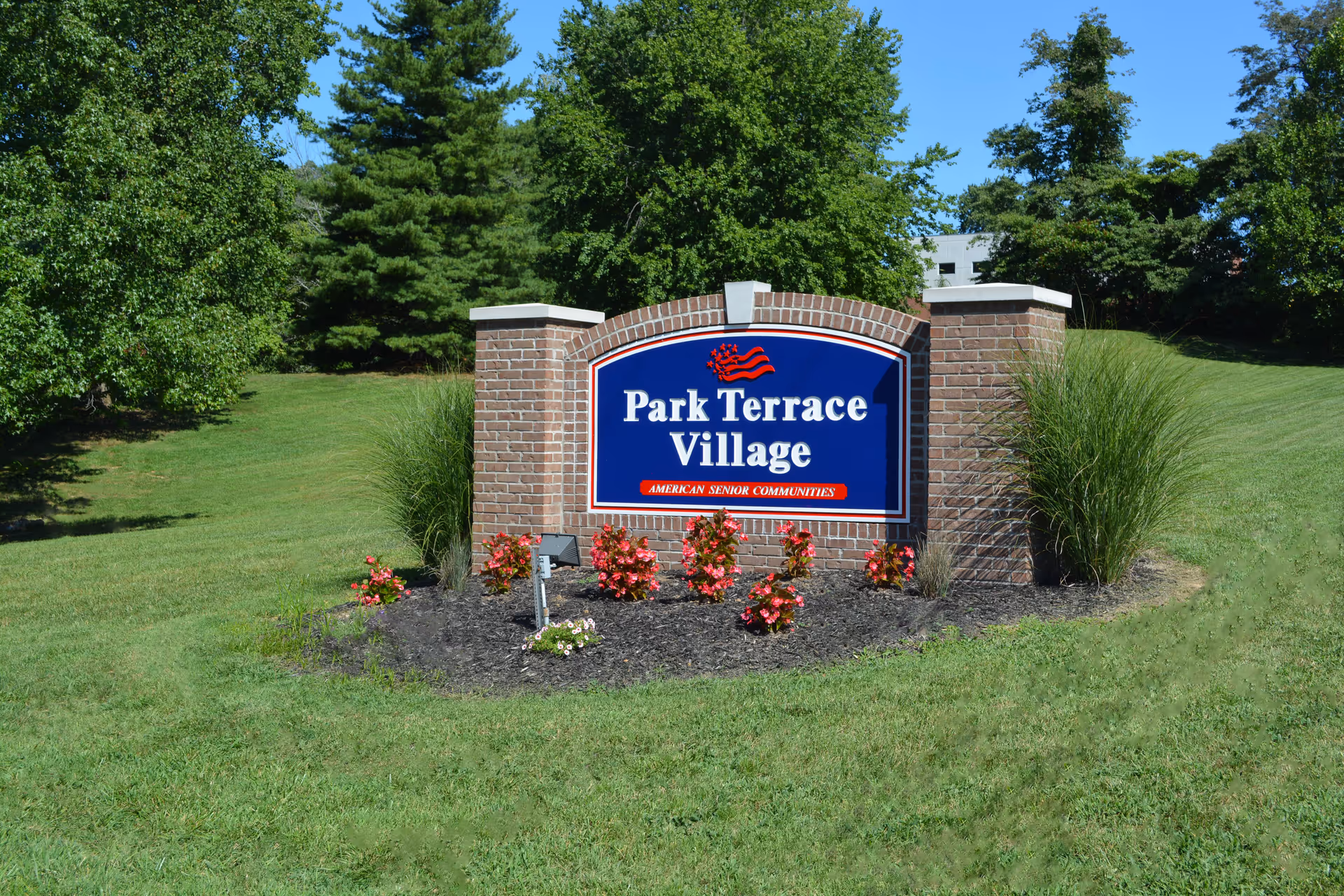 Brick entrance sign reading "Park Terrace Village" on a landscaped grassy lawn with trees behind it.