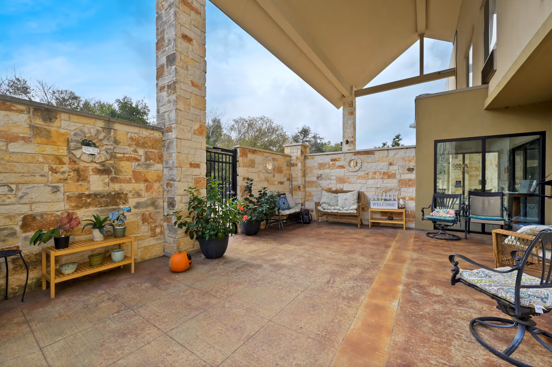 Covered outdoor patio area with stone walls and pillars, featuring various seating options including cushioned chairs and a wicker loveseat. There are potted plants, a small wooden shelf with decorative items, and a pumpkin on the floor. The area is partially enclosed with a gate and has a welcoming atmosphere.