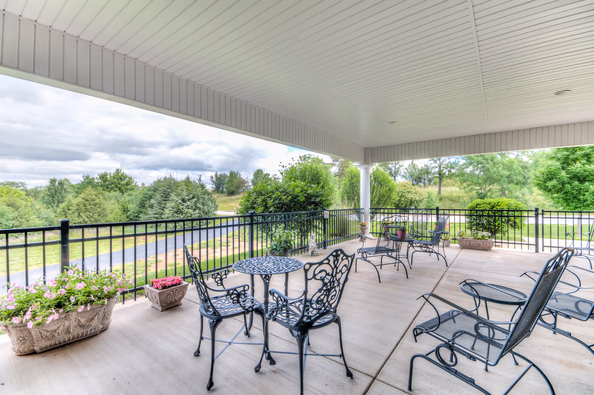 Covered outdoor patio with metal tables and chairs and potted plants overlooking landscaped grounds.