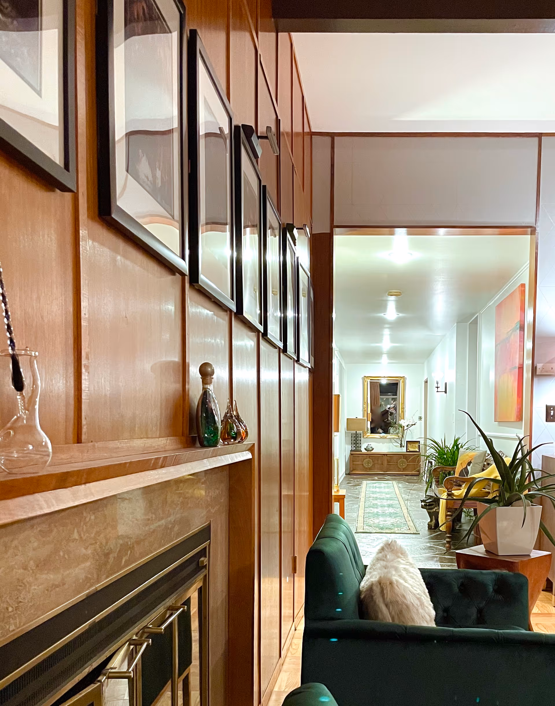Interior view of a living room area with a green tufted sofa featuring a white furry pillow, a wooden paneled wall with framed pictures, a fireplace mantel with decorative glass items, and a hallway leading to another room with plants, a rug, and a large mirror on the far wall.