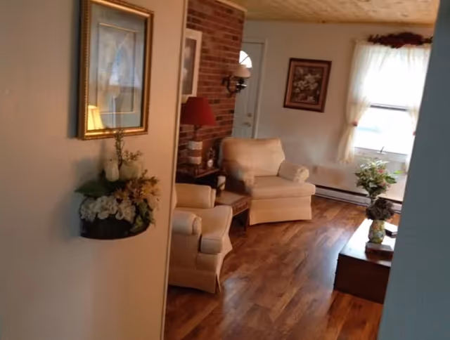 Cozy living room with two beige armchairs, wood floors, a brick accent wall, framed art, and floral arrangements by a sunlit window.