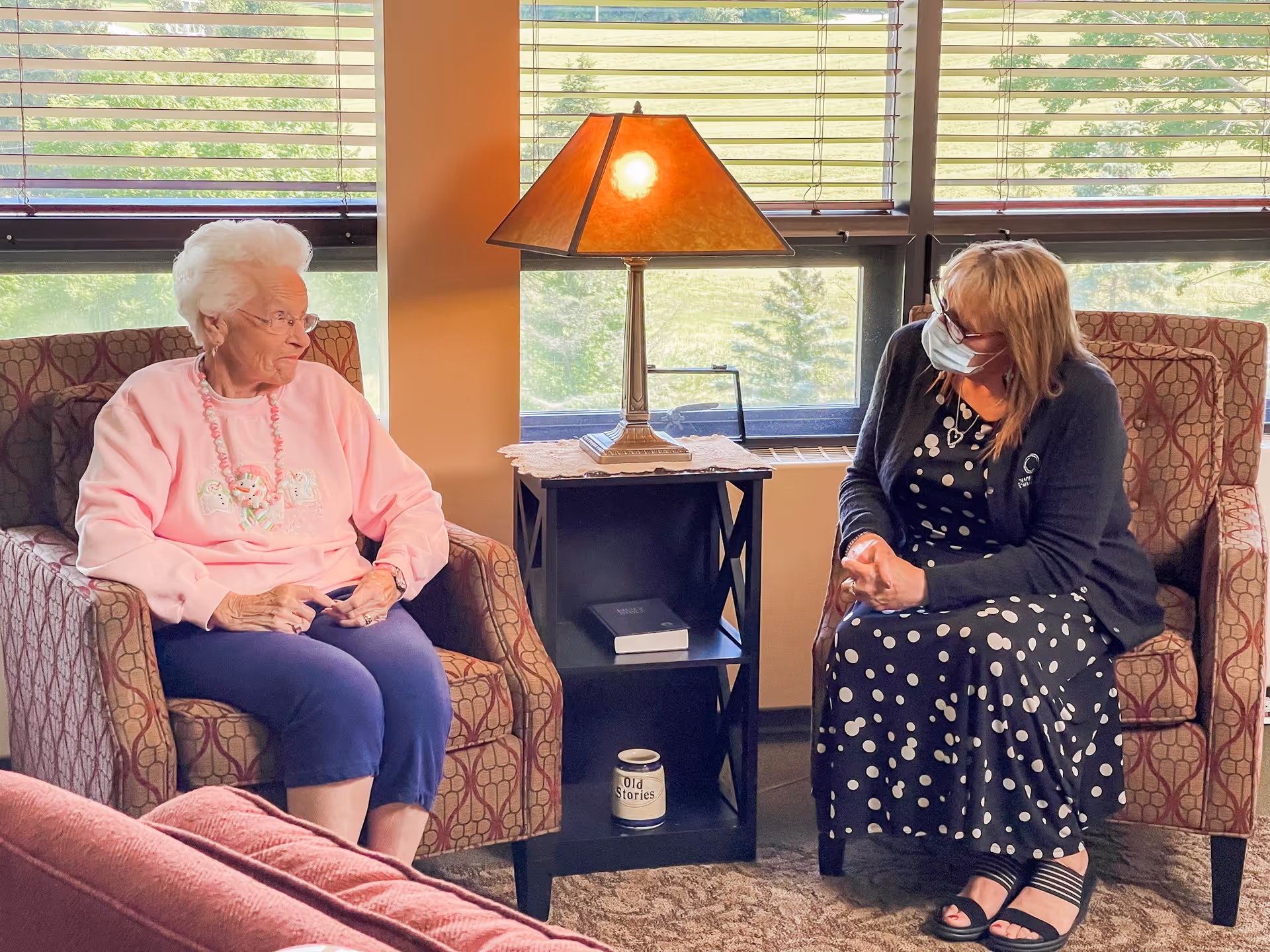 An elderly woman wearing a pink sweater and navy pants sits in a patterned armchair, talking to a woman wearing a black and white polka dot dress, black cardigan, and a face mask, who is seated in a similar armchair. Between them is a small black side table with a lamp on top, a book, and a container labeled 'Old Stories'. Behind them are large windows with blinds partially open, showing greenery outside.