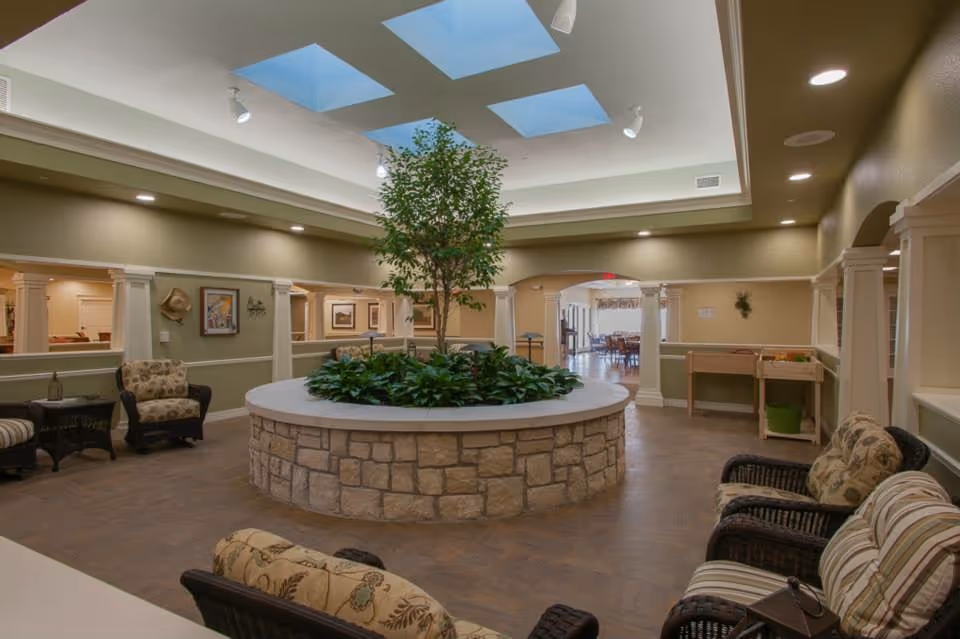 Spacious indoor lounge with a central circular stone planter and tree beneath skylights, surrounded by cushioned chairs and open walkways.