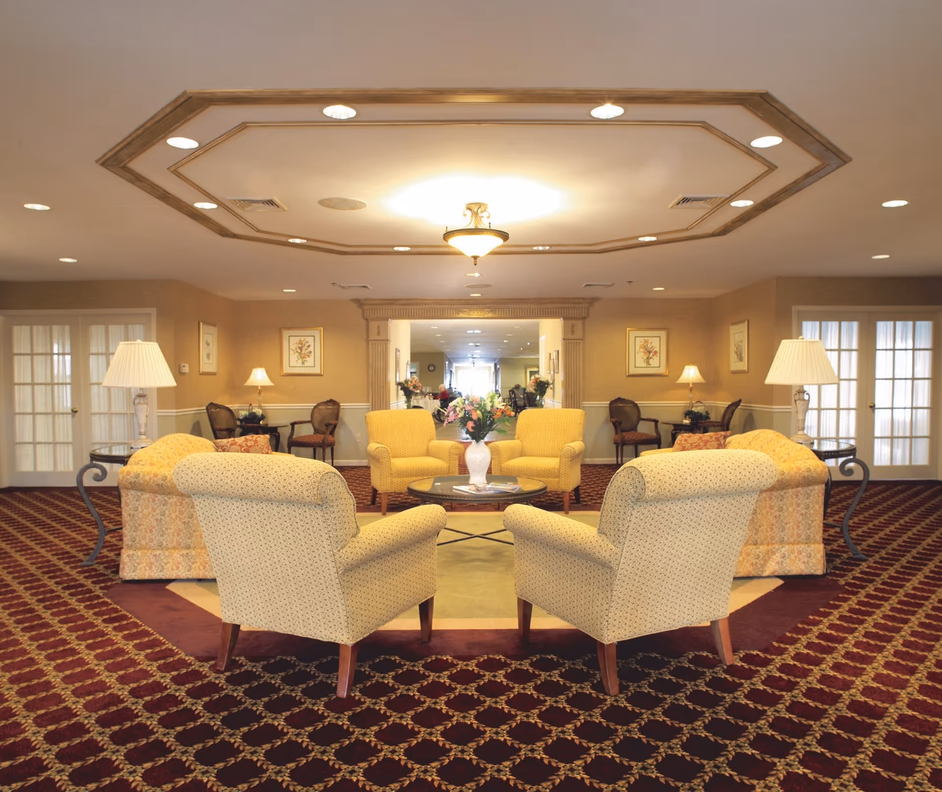 A spacious senior community lounge area with patterned carpet, four upholstered armchairs arranged around a glass coffee table with a floral centerpiece, two side tables with lamps, and additional seating and framed artwork on the walls. The ceiling features recessed lighting and a decorative octagonal design.