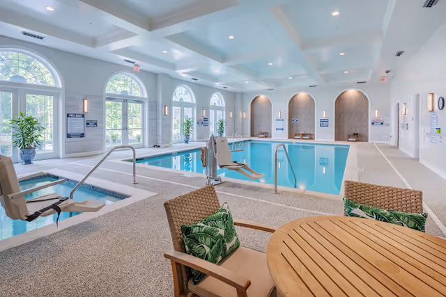 Indoor swimming pool and hot tub area with pool lifts, seating, and arched windows in a bright senior living facility.