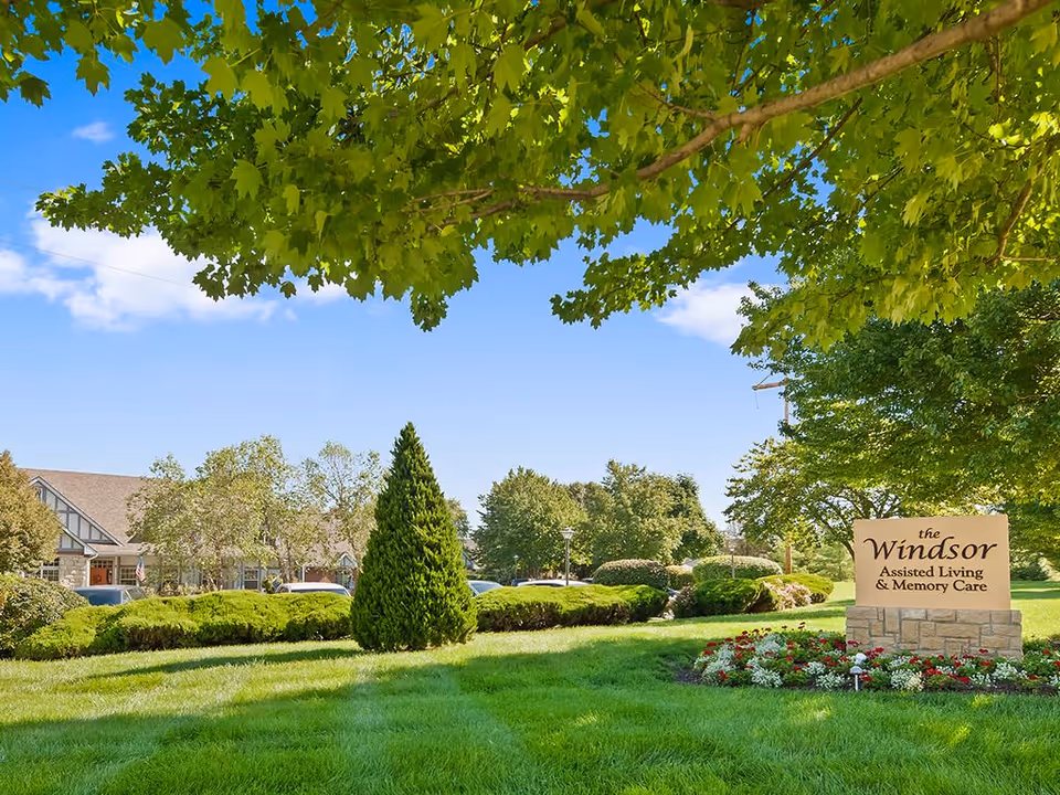 Landscaped lawn and trees with a stone sign reading "The Windsor Assisted Living & Memory Care" and the facility building visible in the background.
