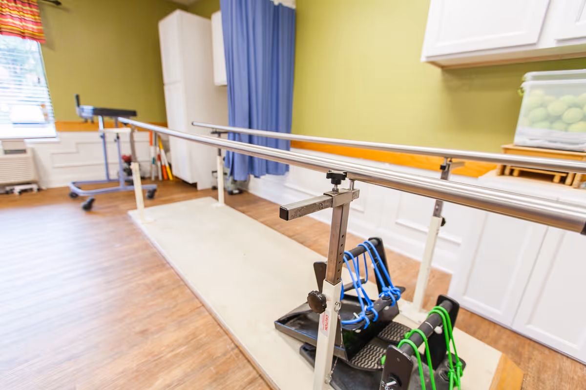 Physical therapy room with parallel bars for walking exercises, resistance bands attached to foot platforms, wooden flooring, green walls, a window with a colorful valance, and medical equipment in the background.