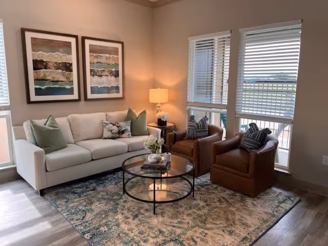 Sunlit living room with a beige sofa, two brown armchairs, round glass coffee table, area rug, lamp, and framed wall art.
