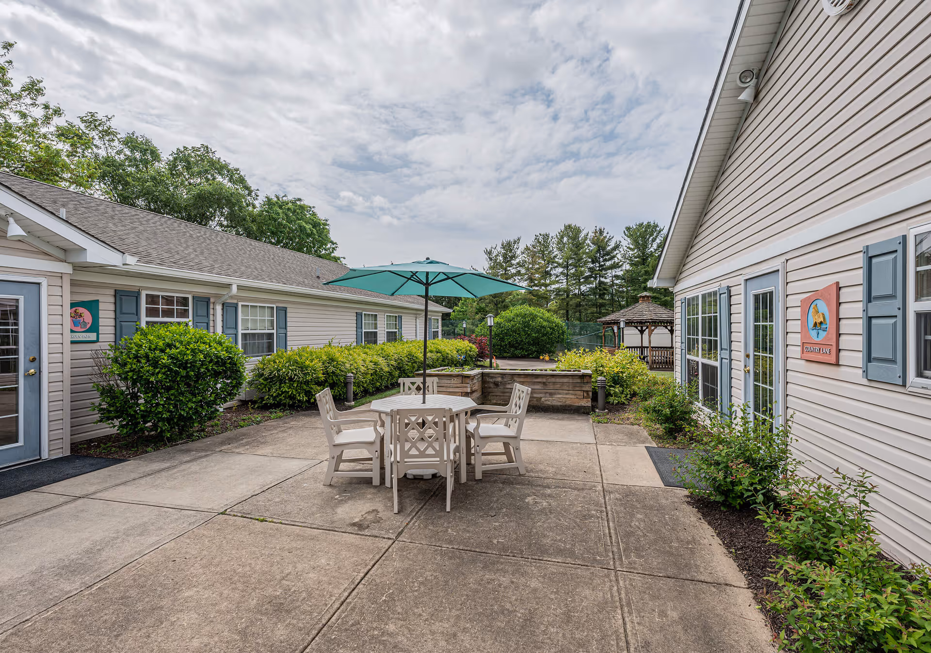Outdoor patio area between two beige buildings with blue shutters, featuring a round table with four chairs and a green umbrella. There are bushes and greenery along the sides, a gazebo in the background, and a partly cloudy sky overhead.