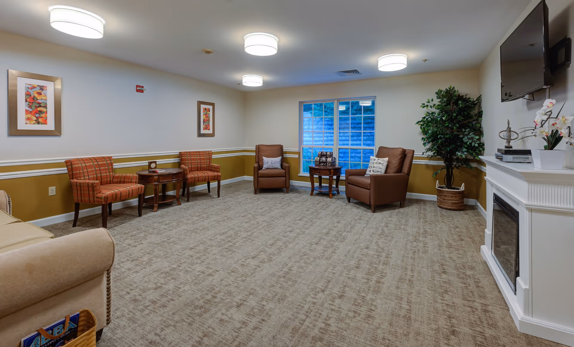 A cozy living room area with beige carpet, two brown leather armchairs with pillows, two red plaid chairs, a beige sofa, two small round wooden tables, a large window, a potted plant, a white fireplace with decorative items, and a wall-mounted TV.